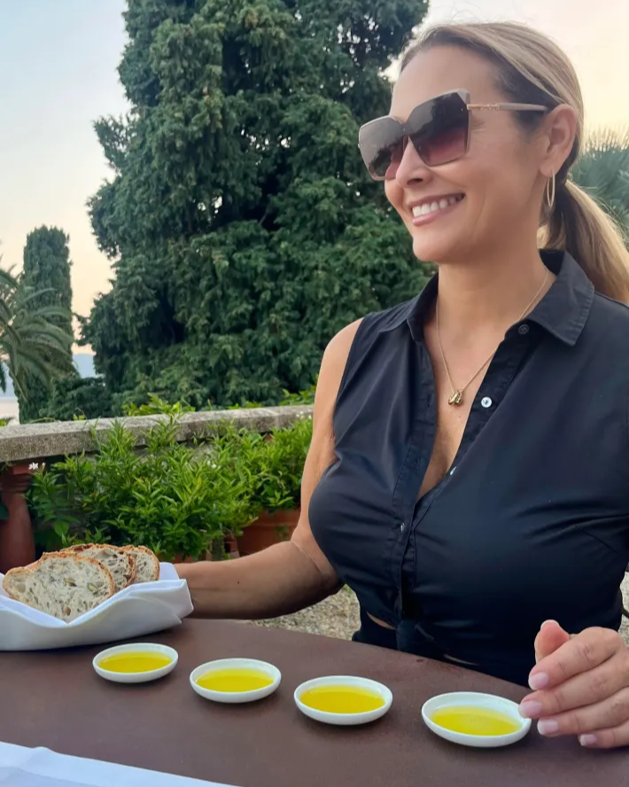 Woman sitting at an outdoor table with olive oil and bread, wearing sunglasses.